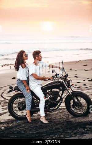 Un couple masculin et féminin vêque de blanc assis sur une moto sur une plage de sable noir à Bali Indonésie sur une moto vintage noire au coucher du soleil Banque D'Images