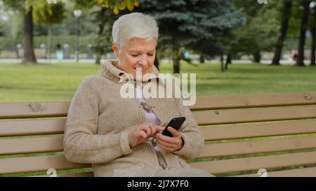 Femme mûre aux cheveux gris surjoyeuse assise à l'extérieur, utilisant le téléphone naviguant dans les applications de smartphone, regardant l'écran, femme sénior discutant avec des parents