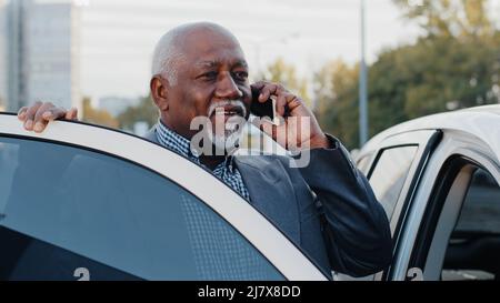 Un homme afro-américain âgé qui parle sur un téléphone portable monte en voiture et s'assoit sur le siège passager avant répond à un appel en utilisant un smartphone mûr Banque D'Images