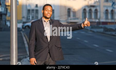 Attrayant étudiant afro-américain, jeune homme en costume attendant le taxi à l'extérieur. Homme d'affaires ou directeur moderne qui fait la gestuelle pour donner un signal au conducteur de voiture Banque D'Images