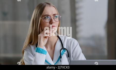 Focalisé caucasien femme pensive infirmière praticien médecin thérapeute en manteau blanc assis à la clinique de l'hôpital pensant à la solution de problème médical Banque D'Images