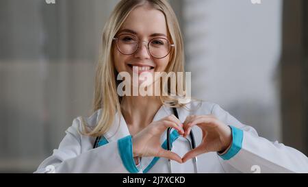 Caucasien médecin spécialiste infirmière femme praticien thérapeute dans les lunettes dans la clinique hôpital à la table montre la forme du coeur signe de miséricorde amour bonté Banque D'Images