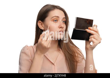 Jeune femme concentrée applique le correcteur tout en regardant dans le miroir. Studio tourné sur fond blanc. Banque D'Images