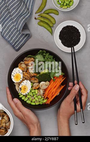 Vue de dessus d'un bol avec nouilles udon japonaises, pétoncles épicés, œufs, carottes, haricots et graines de sésame sur fond de bol noir et de béton gris. Pose à plat Banque D'Images