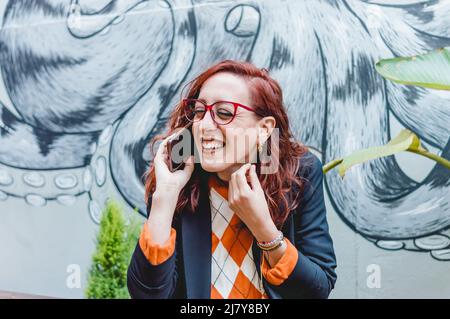 jolie femme rouge caucasienne, portant des vêtements et des lunettes décontractés, en plein air souriant et parlant au téléphone. Banque D'Images