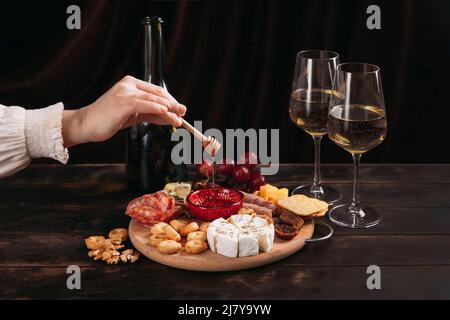 La main de la femme prend un morceau de fromage du tableau des entrées avec un assortiment de fromages, de viande, de rosette à saucisse, de raisin et de biscuits. Banque D'Images