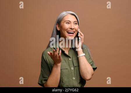 La femme d'âge mûr a une conversation téléphonique agréable. Une femme d'affaires souriante et âgée aux cheveux gris qui parle sur un smartphone isolé sur un fond marron Banque D'Images
