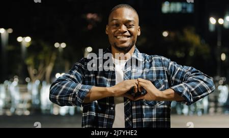 Homme afro-américain homme debout dans la ville de nuit affectueux portrait romantique regardant l'appareil-photo faire le coeur avec les doigts. Beau garçon Banque D'Images