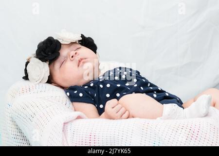 belle petite fille de latina reposant sur un drap blanc, vêtue d'un costume bleu de barboteuse, de bas blancs et d'un bandeau de fleurs noir et blanc. maternité c Banque D'Images