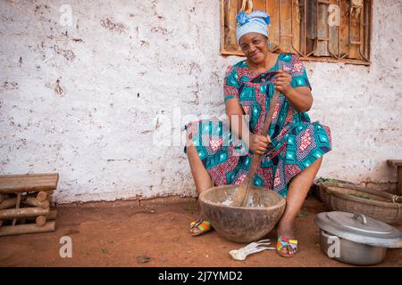 Une femme africaine cuisine avec un mortier et un pilon, elle est habillée dans une robe traditionnelle et elle est dans le village Banque D'Images