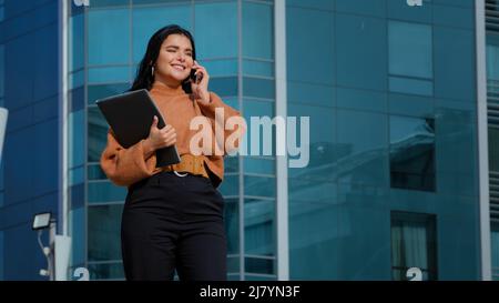 Une femme d'affaires heureuse se tient sur le bureau en arrière-plan parle sur le téléphone mobile répond à un appel convivial communique avec un ami en utilisant les dépenses de smartphone Banque D'Images