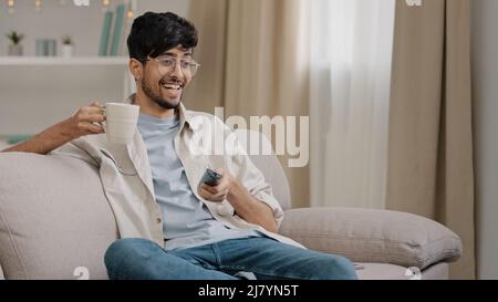 Homme arabe barbu gars avec des lunettes de repos à la maison assis sur le canapé boire thé café regarder un film drôle championnat de sport jeu de télévision se réjouit à Banque D'Images
