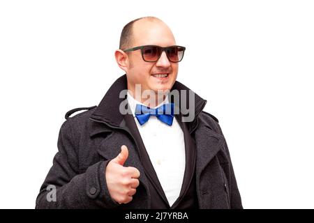 Un homme bald adulte portant des lunettes de soleil et vêtu de vêtements élégants et d'un noeud papillon bleu tient un de ses pouces tout en souriant. Le mâle est sur un wh Banque D'Images