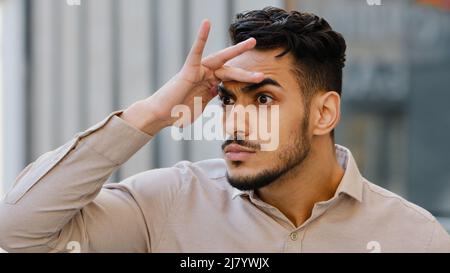 Gros plan drôle humoristique barbu homme non rasé hispanique arabe Indien gars debout dans la rue à l'extérieur de la ville tenant la paume des yeux à la recherche de quelqu'un Banque D'Images