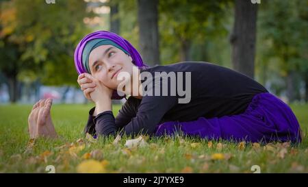 Belle jeune femme islamique porte hijab assis sur l'herbe dans le parc à l'extérieur se pliant dans le corps ventre étirement faisant l'exercice de yoga physique Banque D'Images