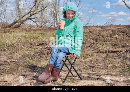 Une petite fille de race blanche heureuse positive dans une veste verte est assise sur une chaise pliante de l'eau potable d'un verre à un pique-nique au printemps. Banque D'Images