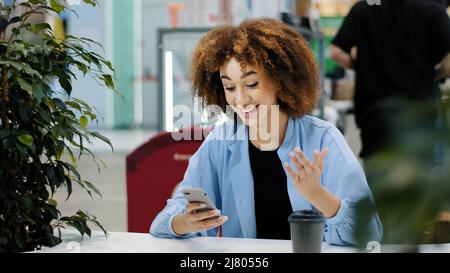 Jeune fille adolescente millénaire afro-américaine avec des cheveux bouclés assis dans un café regardant le téléphone mobile se réjouir gagne la victoire bonne Banque D'Images