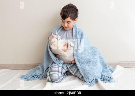 Un garçon en pyjama gris est placé sur le sol sur une couverture bleue tricotée, avec un beau chat blanc, Banque D'Images