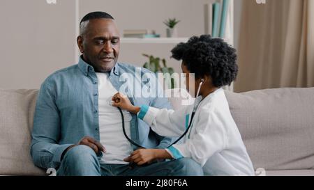 Petite fille africaine américaine dans la robe médicale joue assis sur le canapé enfant médecin écoute le père stéthoscope tend l'infirmière ayant plaisir aimer fille Banque D'Images