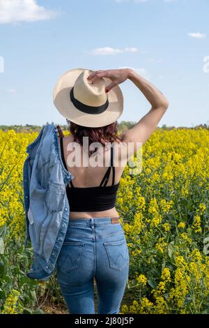 Vue arrière d'une femme debout dans un champ de colza au printemps avec sa main sur la tête, France Banque D'Images