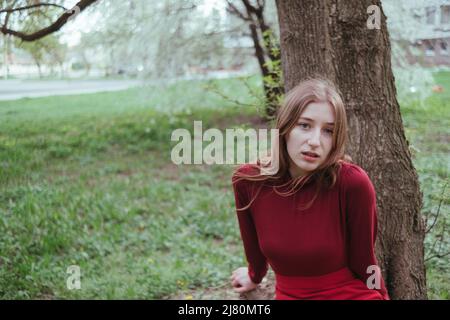 une fille en rouge penche sur un arbre et regarde mystérieusement dans l'appareil photo Banque D'Images