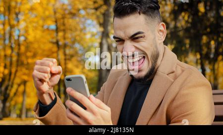 Un homme hispanique enthousiaste lisant de bonnes nouvelles sur un smartphone assis sur un banc dans le parc d'automne jeune homme heureux surpris de recevoir un message trop joyeux homme Banque D'Images