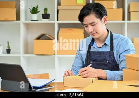 Un jeune homme asiatique travaillant dur empaquemet un produit de livraison, en écrivant le nom et l'adresse d'un client sur une boîte à colis dans la salle de stockage. PME, commerce électronique Banque D'Images