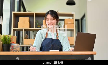 Charmante femme asiatique du millénaire planifiant sa liste de matières premières à commander avec son fournisseur, travaillant sur ordinateur portable. Entreprise de démarrage Banque D'Images