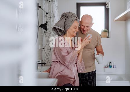 Couple senior amoureux dans la salle de bains, utilisant un smartphone, concept de routine du matin. Banque D'Images