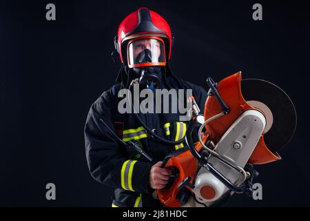 portrait d'un pompier puissant en uniforme résistant au feu tenant une tronçonneuse à hache dans ses mains, studio à arrière-plan noir Banque D'Images