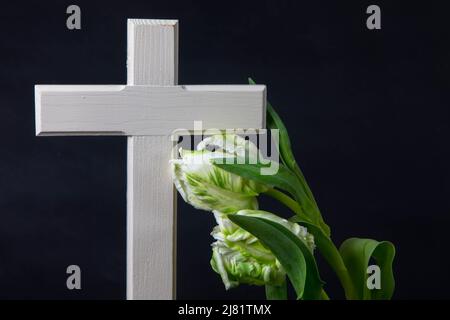 Croix religieuse en bois avec fleurs de tulipe blanches sur fond sombre pour avis nécrologique. Moment de chagrin à la fin d'une vie. Dernier adieu. Banque D'Images