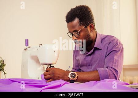 portrait d'un beau homme africain souriant couturière avec machine à coudre travaillant dans un atelier sur mesure mannequin, mètre ruban de table dans la pièce contre Banque D'Images