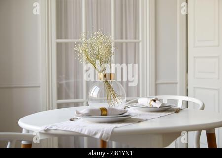 Vase en verre élégant avec fleurs douces et assiettes en céramique avec serviettes roulées servies sur une table ronde dans la salle à manger blanche Banque D'Images