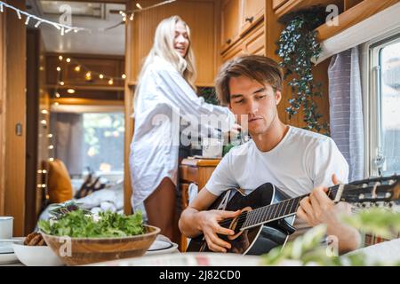 Homme jouant de la guitare, chant sur une petite cuisine, cuisine en van. Jeune fille, gars juste couple marié, amoureux voyageant en camping-car, maison sur roues, traile Banque D'Images