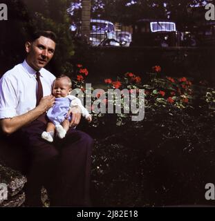 Un père est assis avec son bébé fils dans un jardin à Corton, Suffolk, en 1957. Le père porte une chemise blanche et une cravate sombre. 1950s voitures sont visibles derrière les garde-corps du jardin. Banque D'Images