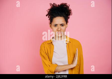 Triste maudit fille afro-américaine dans des vêtements décontractés, en disant non, montre stop geste avec la paume sur le côté, exprimant l'insatisfaction, debout sur un arrière-plan isolé de couleur rose, regardant l'appareil photo Banque D'Images