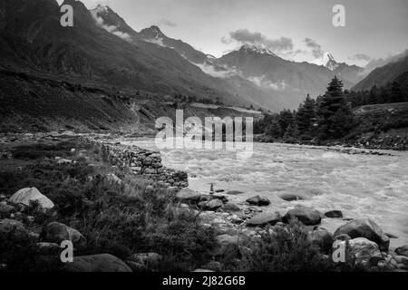 La rivière Baspa flanquée de rochers, d'arbres et de l'Himalaya avec des sommets enveloppés dans des nuages sous ciel couvert en été à Chitkul, en Inde. Banque D'Images