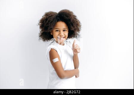 Une petite fille afro-américaine positive, préchooler, avec un bracelet d'aide sur l'épaule après la vaccination, pour l'immunité contre covid19, et d'autres maladies, se tiennent sur fond blanc isolé, mouvement de pouce vers le haut Banque D'Images