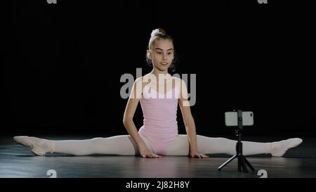 Jeune fille adolescent ballerina acrobat gymnaste s'assoit sur le plancher cours de danse en ligne avec l'entraîneur d'Internet professeur à distance avec smartphone sur Banque D'Images