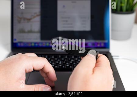 Russie, Krasnodar - 15 avril 2022 : homme travaillant sur un ordinateur portable et utilisant un portefeuille matériel de crypto-monnaie. Solution de stockage sécurisée pour bitcoin. Banque D'Images