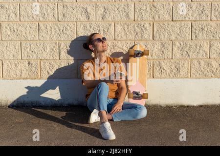 Jeune skate décontracté assis souriant et heureux de profiter de la chaude source de respiration sifflante avec son skateboard incliné sur le mur Banque D'Images