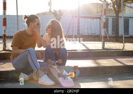 Mignon jeune couple d'adolescents s'amusant et plaisantant avec le skate-board autour au coucher du soleil avec des fusées autour en été Banque D'Images