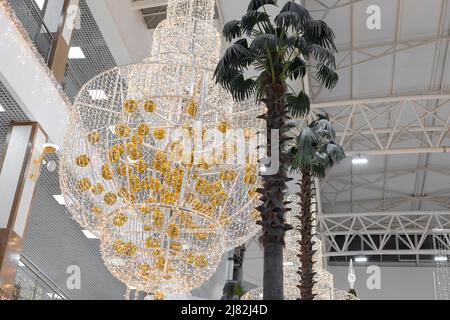 Un immense lustre décoré avec soin avec des guirlandes et des boules dorées du nouvel an est suspendu dans le hall du centre commercial au niveau de grands palmiers. Banque D'Images