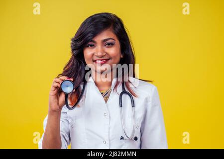 Femme indienne jeune et belle blonde femme gynécologue médecin utilisant le stéthoscope dans un manteau médical blanc sur un fond jaune dans le studio Banque D'Images