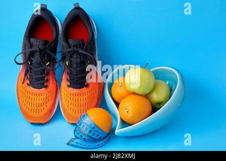Des baskets orange, un mètre ruban et un bol en forme de coeur avec des pommes de fruits et des oranges sur fond bleu. Equipement amincissant Banque D'Images