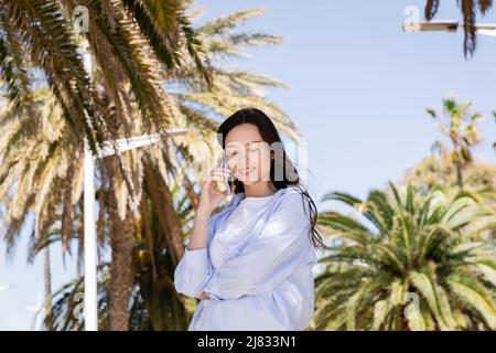 brunette femme en chemise bleue souriant en parlant au téléphone portable dans un parc urbain Banque D'Images