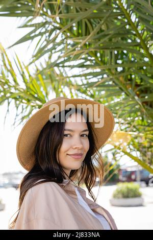 portrait d'une femme brune en chapeau de paille souriant à l'appareil photo près d'un palmier flou Banque D'Images