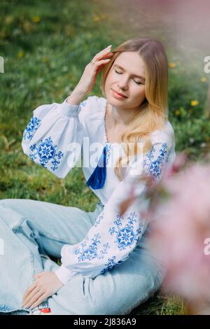 Jeune femme blonde repose sur la pelouse dans le jardin vêtu d'une chemise ukrainienne brodée près d'un arbre sakura en fleurs. Gros plan. Banque D'Images