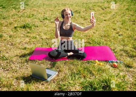 Bonne blonde cheveux sport fille écoute de la musique et prend un selfie, pendant le yoga sur le tapis au parc de la ville en été Banque D'Images