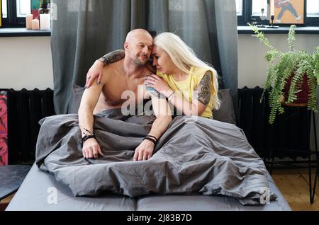 Tatouée femme caucasienne mature avec de longs cheveux gris assis sur le lit le matin embrassant son petit ami Banque D'Images